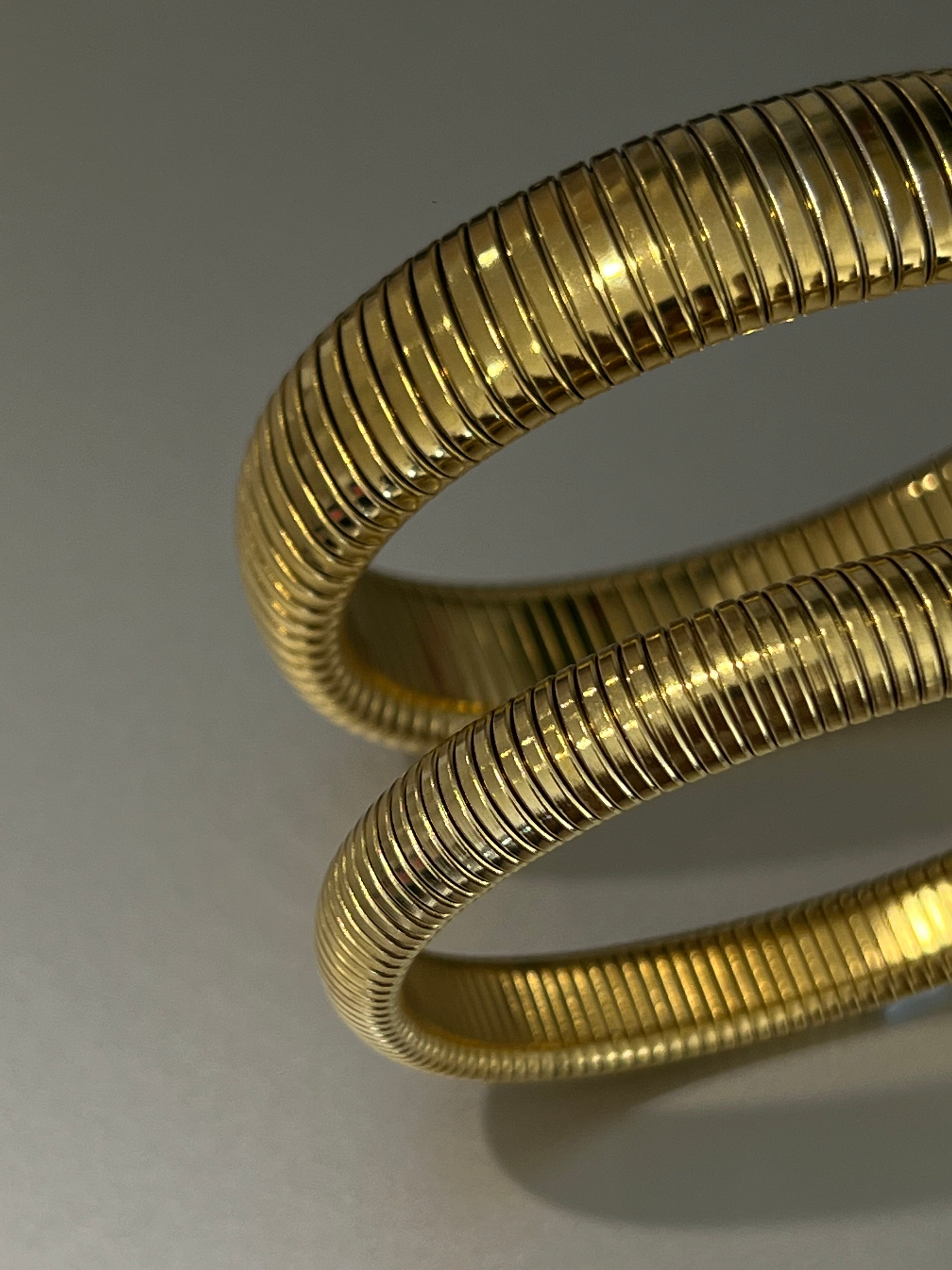 Close-up of two coiled gold EverLuxe cuff bracelets stacked against a dark fabric backdrop.