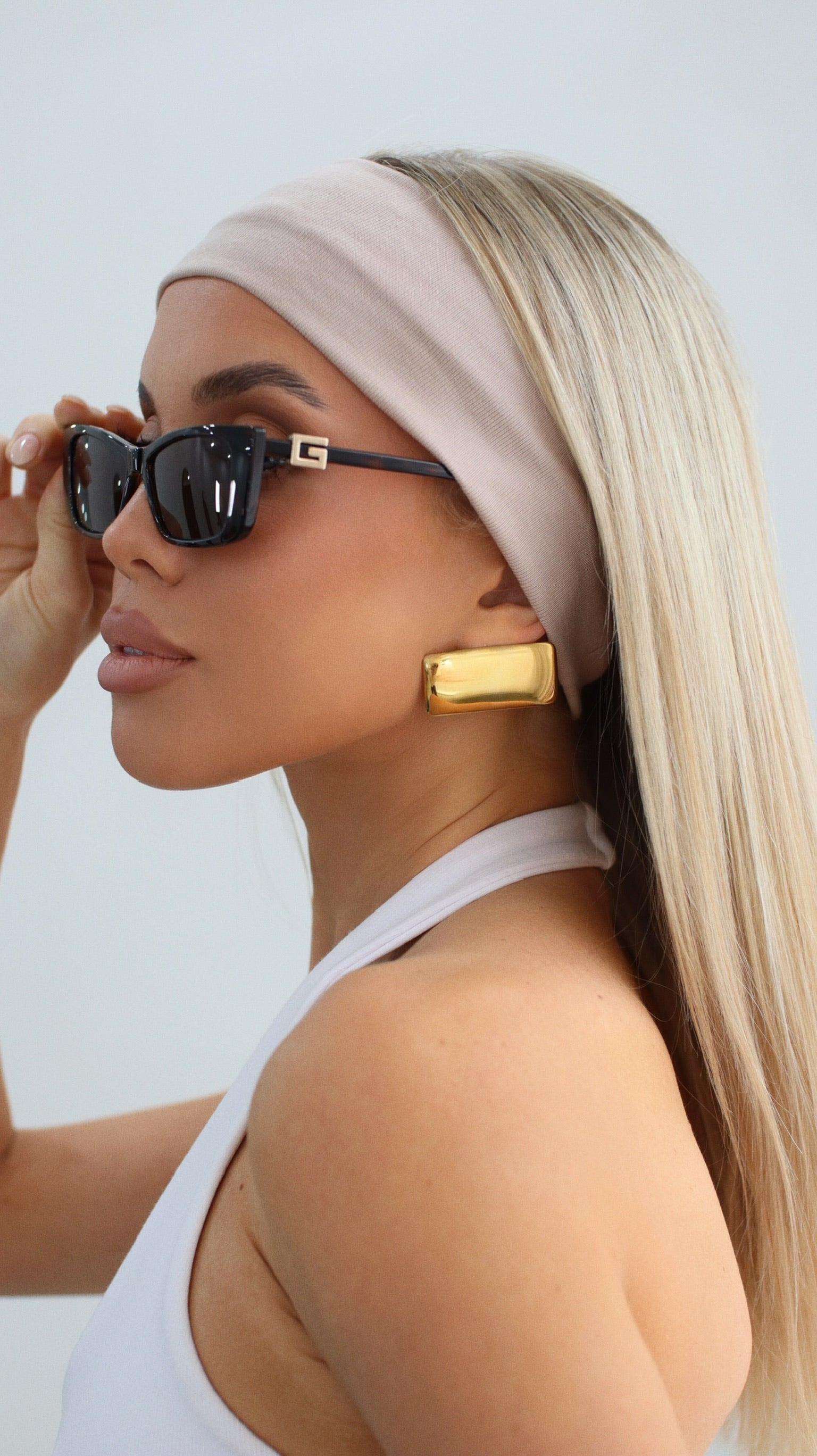 Blonde woman in sunglasses and a white headband wearing chunky gold rectangular earrings, looking to the side.
