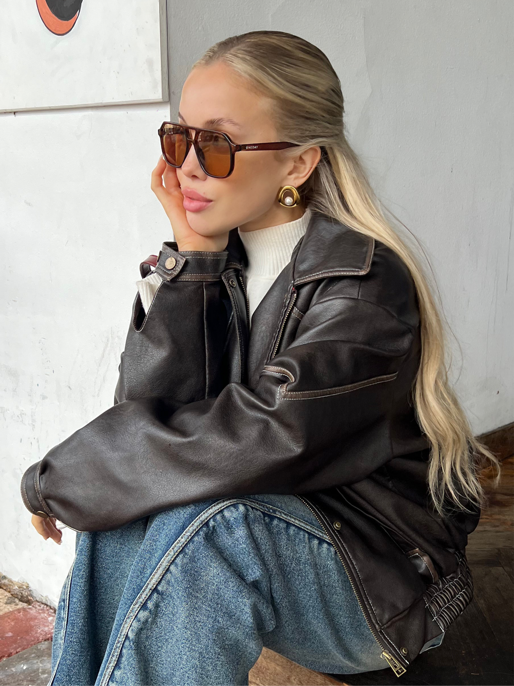 Woman wearing Sol Halo gold earrings, a black leather jacket and sunglasses against a white wall.