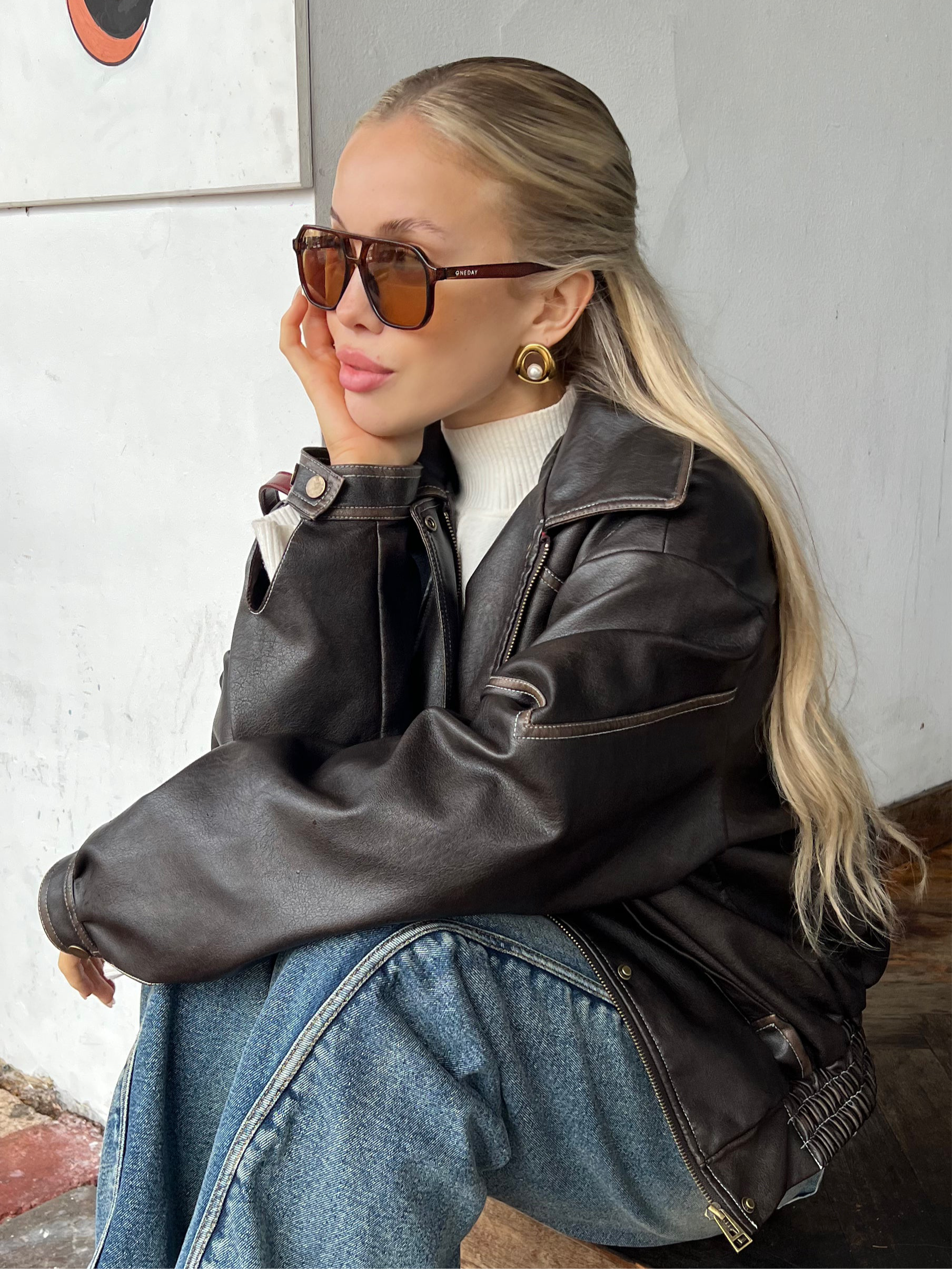 Woman wearing Sol Halo gold earrings, a black leather jacket and sunglasses against a white wall.