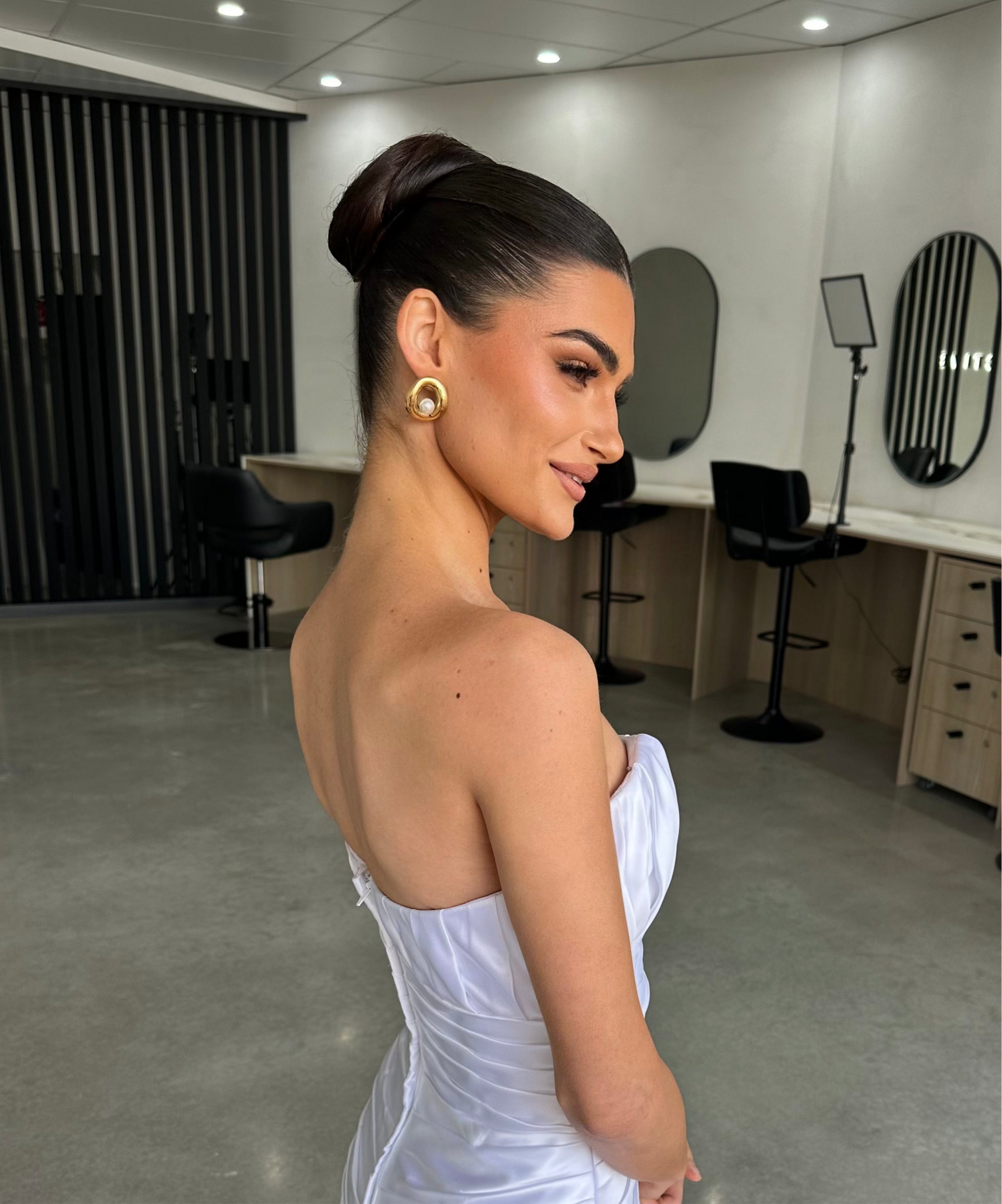 Woman in a white strapless dress posing in a room with mirrors and chairs. she is wearing everluxe earrings.