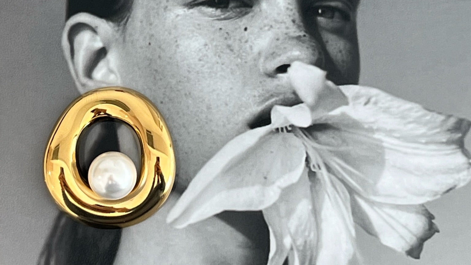 Person wearing a gold earring with a pearl, holding a white flower to their nose against a gray background